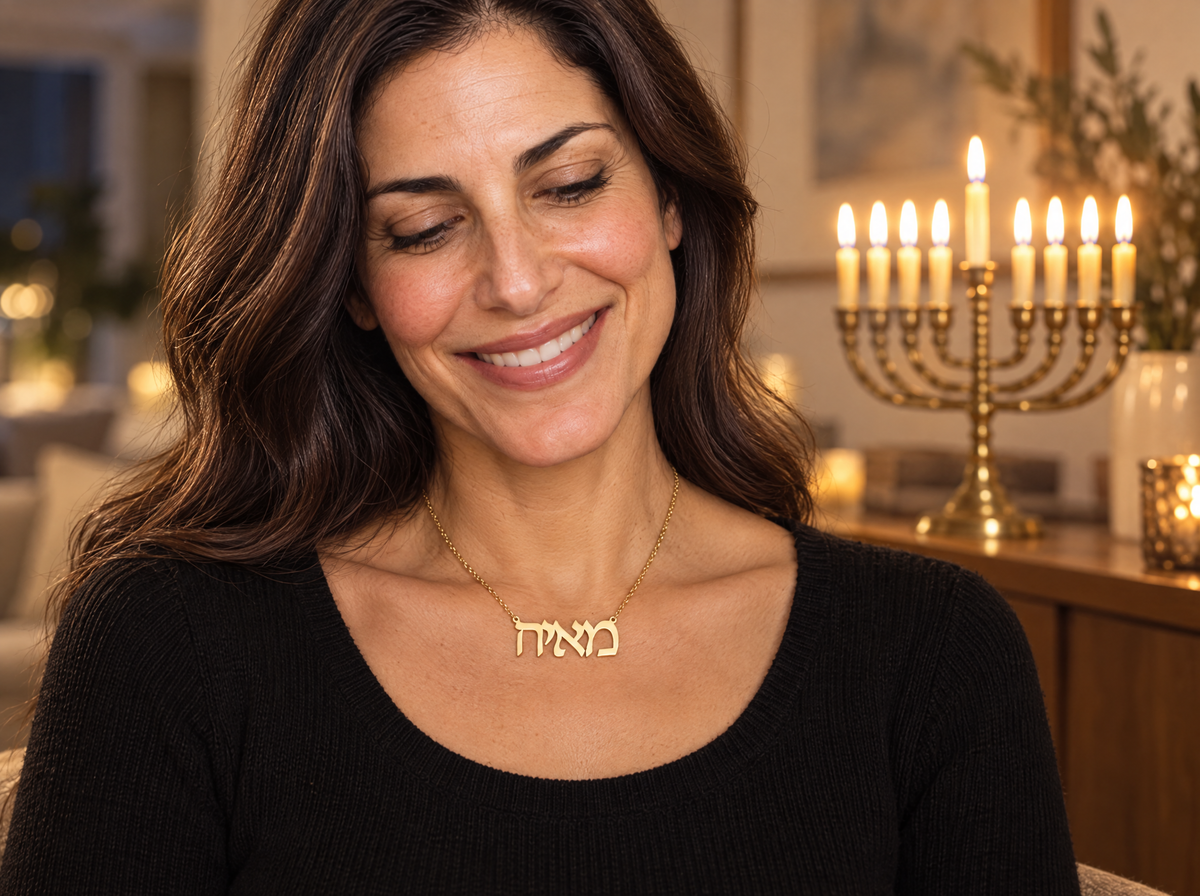Smiling woman wearing a Hebrew name necklace reading מאיה in 18K gold plating, with a lit Hanukkah menorah behind her