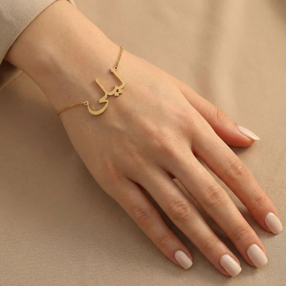 Gold Arabic name bracelet worn on a fair hand with clean natural nails, resting on a smooth neutral beige surface with minimal elegant styling.