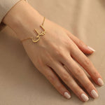 Gold Arabic name bracelet worn on a fair hand with clean natural nails, resting on a smooth neutral beige surface with minimal elegant styling.