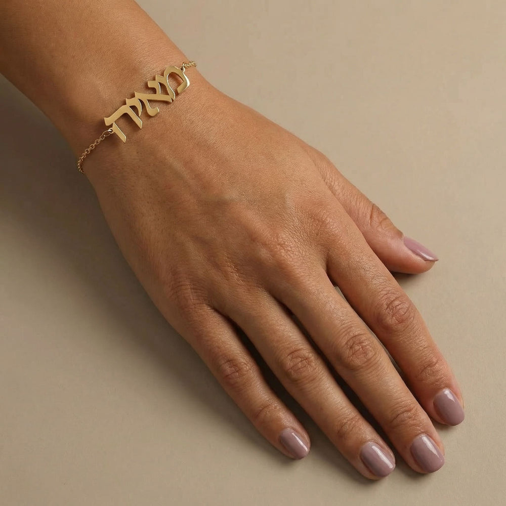 Gold Hebrew name bracelet worn on a lightly tanned wrist, hand relaxed with soft pink nails, captured on a warm cream background in diffused light.