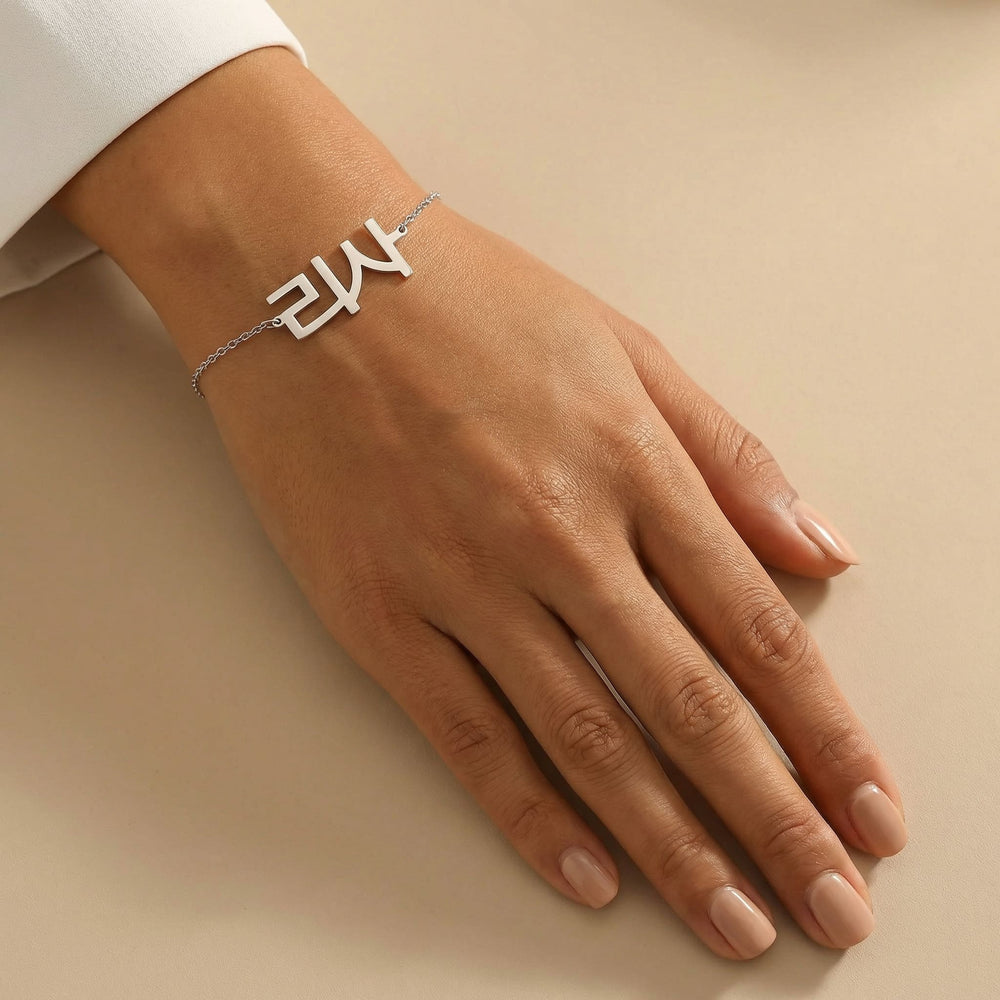 Silver Korean name bracelet displayed on a tanned wrist with clean unpolished nails, styled on a smooth cream background under natural daylight.
