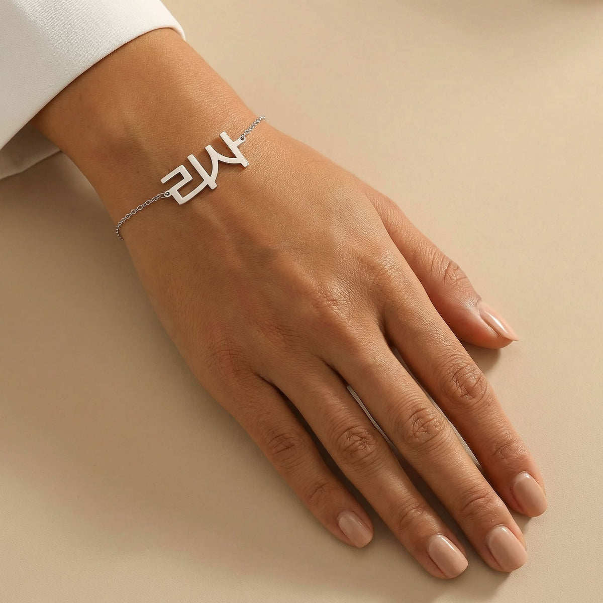 Silver Korean name bracelet displayed on a tanned wrist with clean unpolished nails, styled on a smooth cream background under natural daylight.