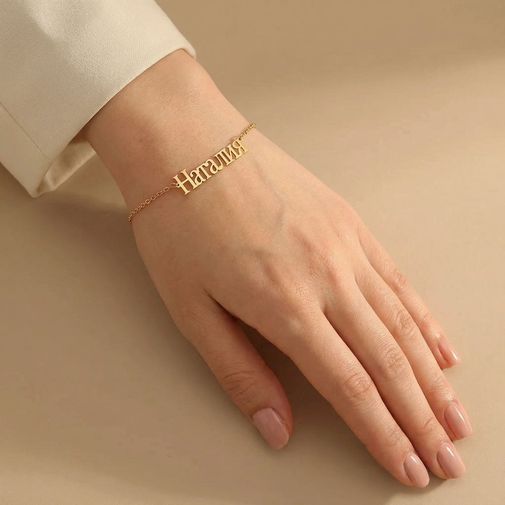 Gold Russian name bracelet worn on a fair toned hand with light pink manicured nails, placed against a warm beige background with soft shadows.