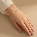 Gold Russian name bracelet worn on a fair toned hand with light pink manicured nails, placed against a warm beige background with soft shadows.