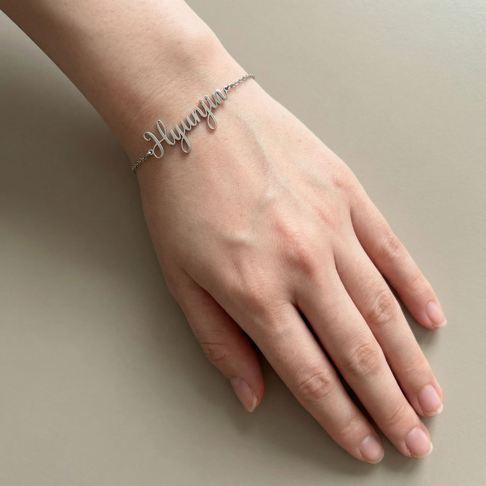 Silver Kpop idol name bracelet reading Hyunjin worn on a fair wrist, hand relaxed with natural nails, photographed on a neutral beige background.