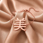 Rose gold necklace with a Kanji character pendant meaning "beauty" on a beige fabric background