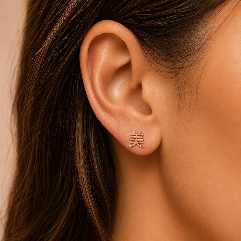Close-up of an ear wearing a rose gold earring with a Kanji character meaning "beauty", against a neutral background.