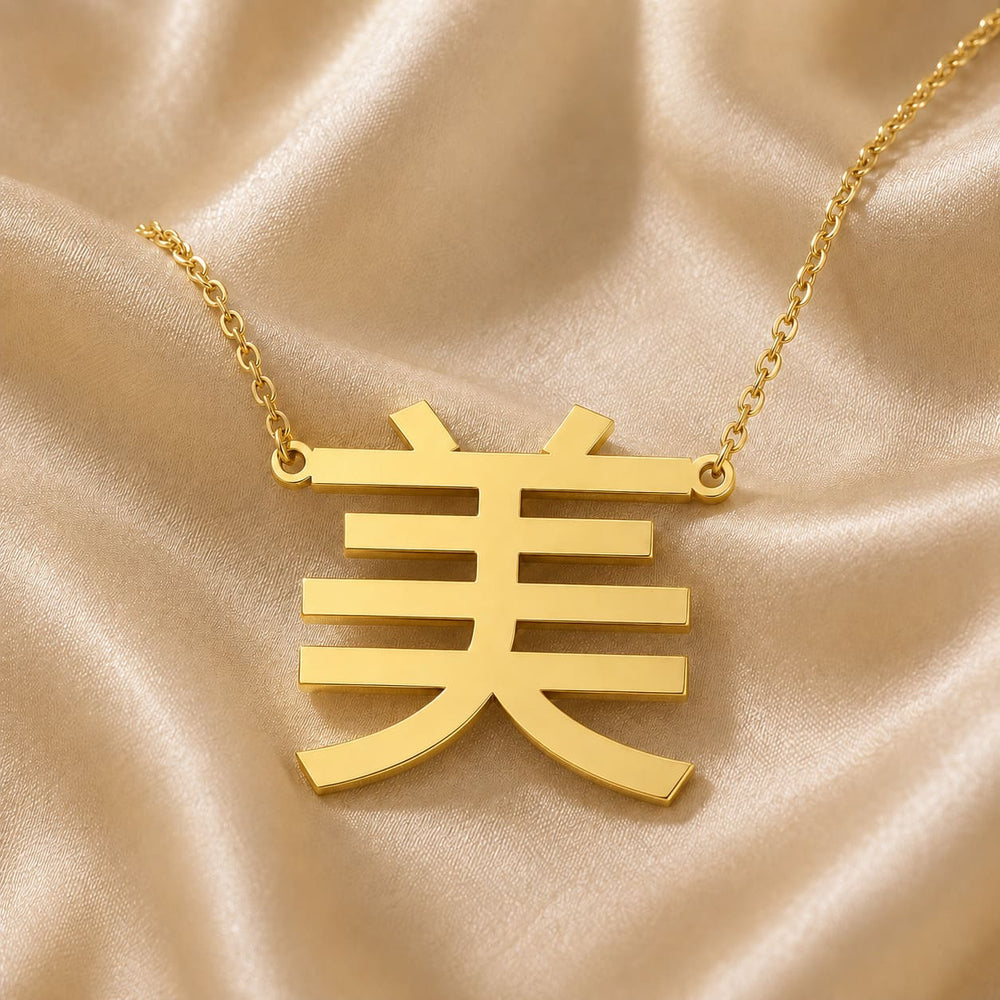 Gold necklace with a Kanji character pendant meaning "beauty" on a beige fabric background