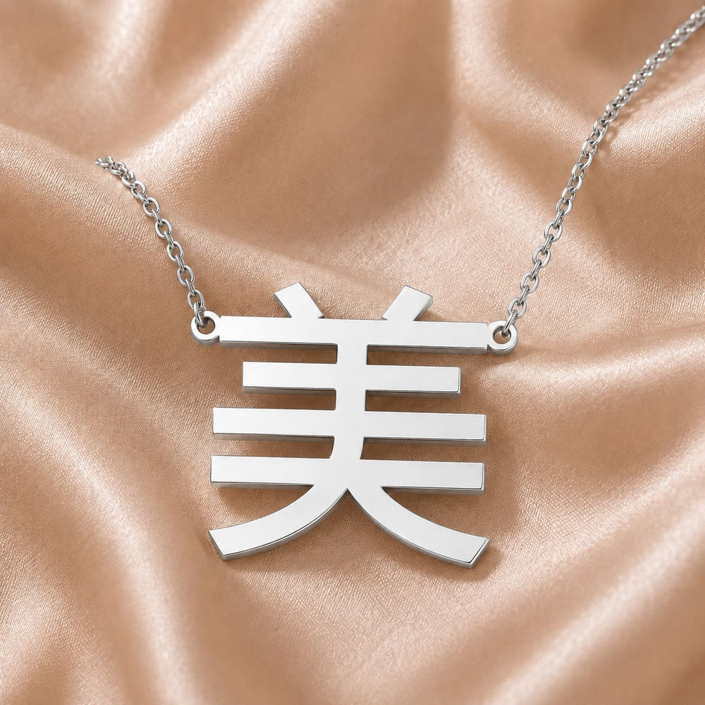 Silver necklace with a pendant featuring a Kanji character meaning "beauty" on a beige fabric background