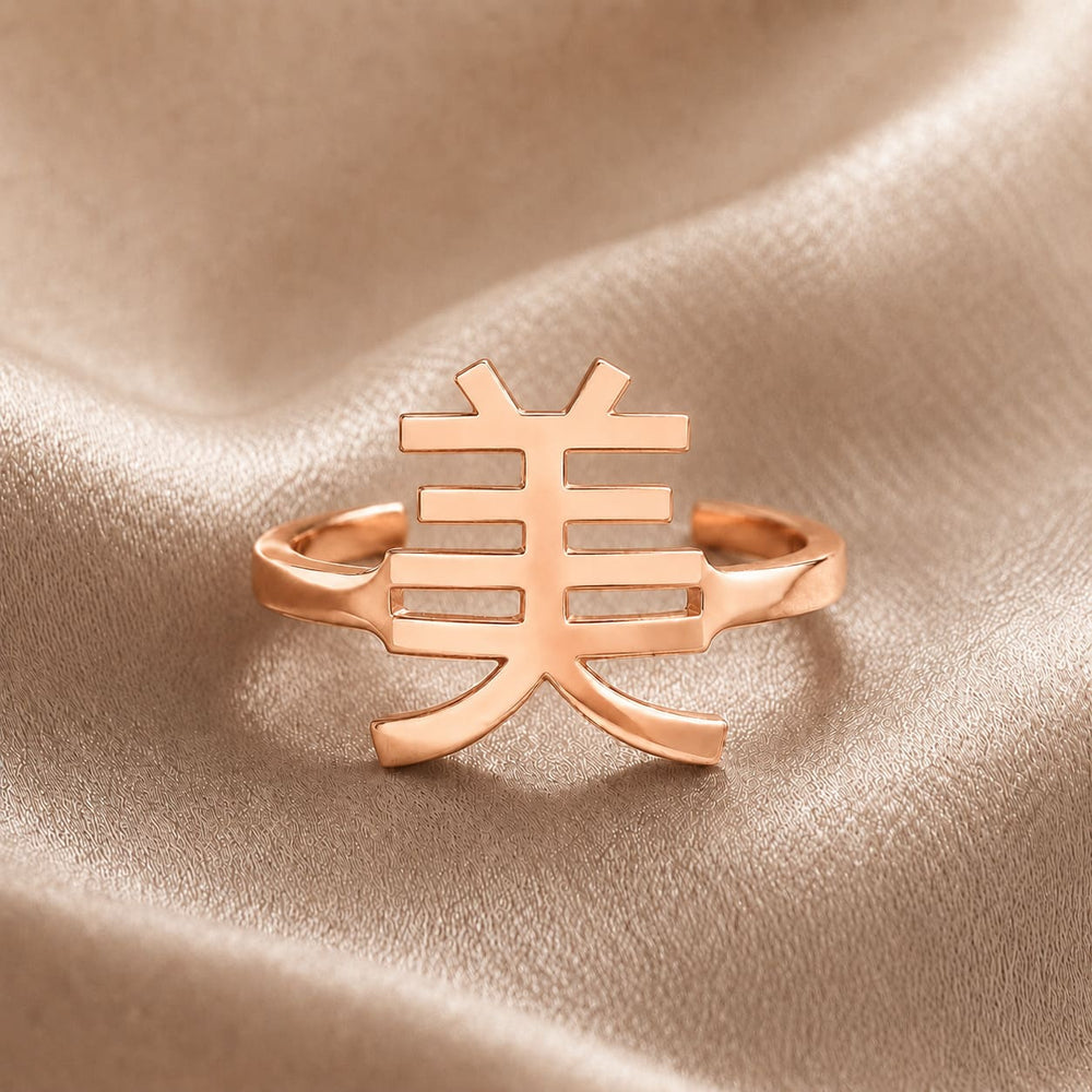 Rose gold ring with a Kanji character pendant meaning "beauty" on a beige fabric background