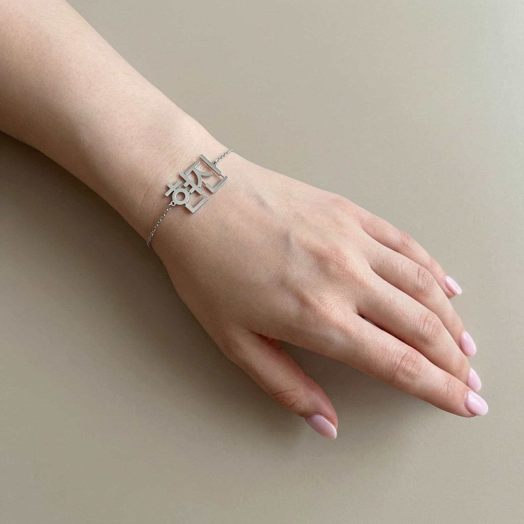 Silver Korean Hangul name bracelet worn on a fair toned wrist, shown on a relaxed hand with natural short nails against a soft beige background in natural light.