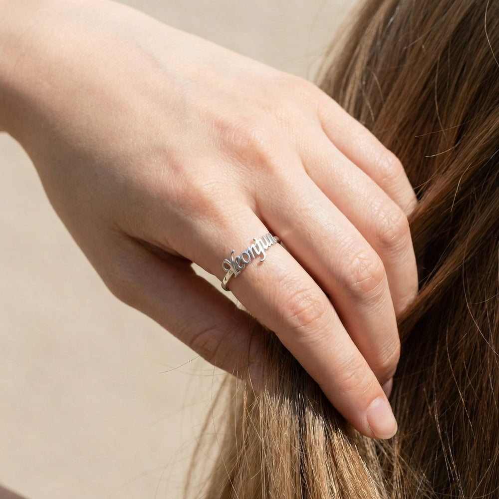 Woman's hand wearing a silver custom name ring with "Yeonjun" in cursive script while gently playing with brown hair, showing the ring on the middle finger in a casual lifestyle pose.