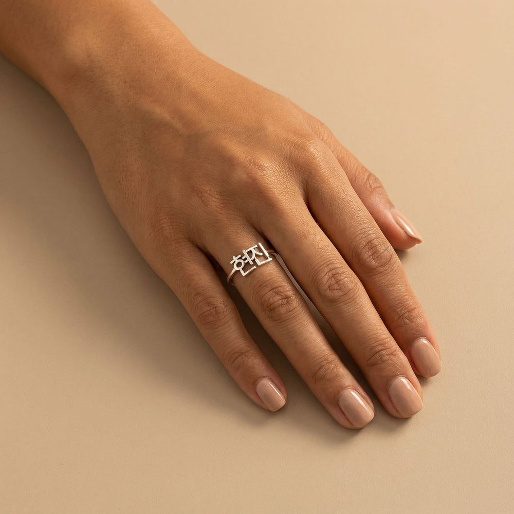 Woman's hand wearing a silver custom Korean Hangul name ring with "현진" (Hyunjin) on the middle finger, hand flat on plain beige background with nude nails.