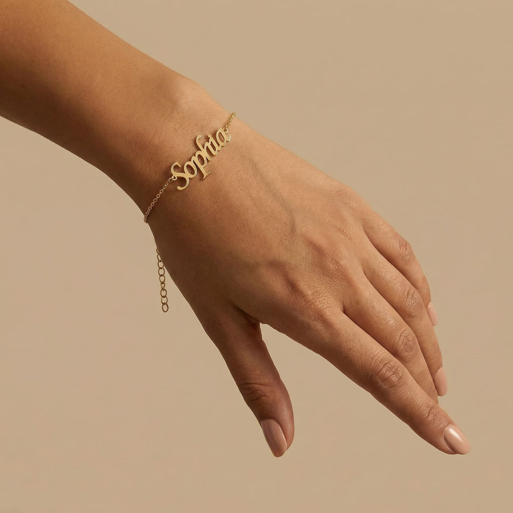 Gold Latin name bracelet reading Sophia worn on a medium toned hand with nude polished nails, photographed on a warm cream background with soft shadows.
