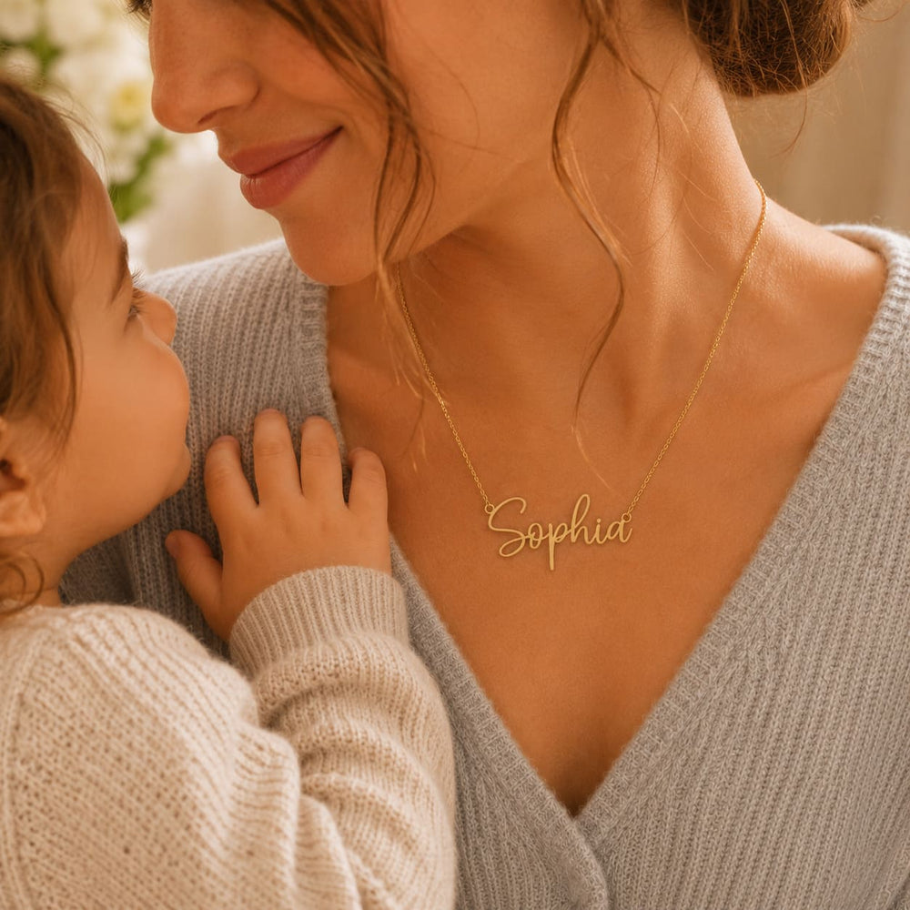 Mother wearing a gold 'Sophia' custom name necklace while holding her child in a warm lifestyle moment while looking into each others eyes