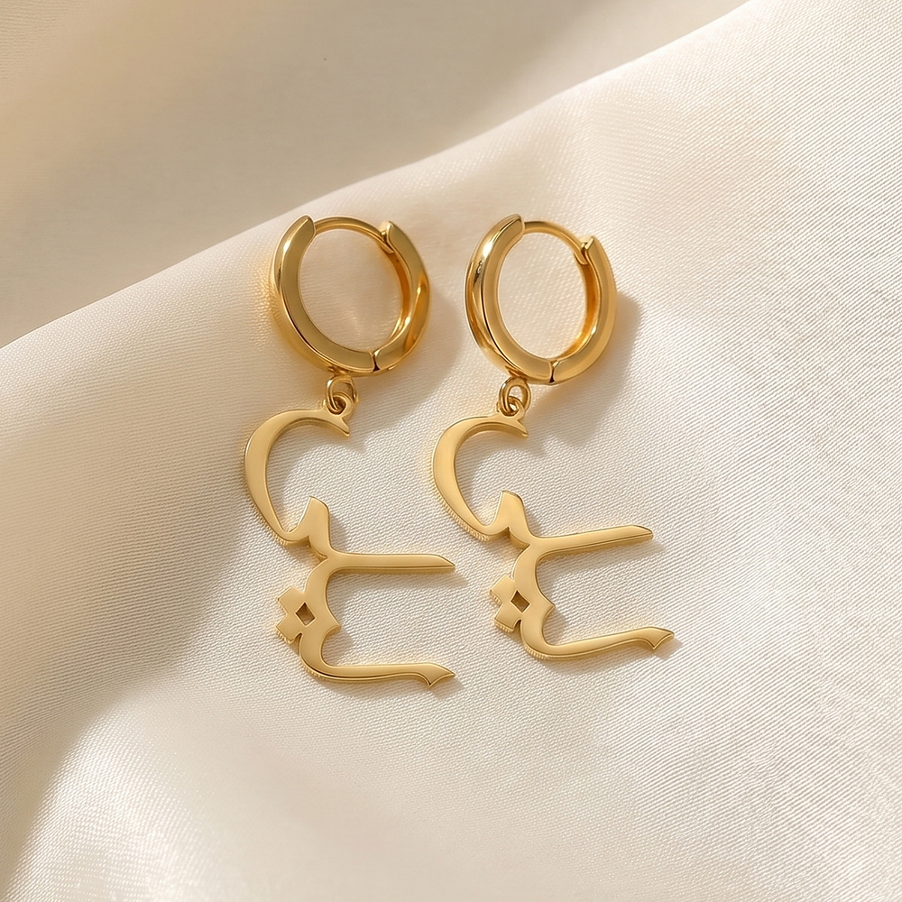 Custom Arabic name drop earrings in 18K gold plating, a pair of huggie hoops with Arabic name charms, on cream silk