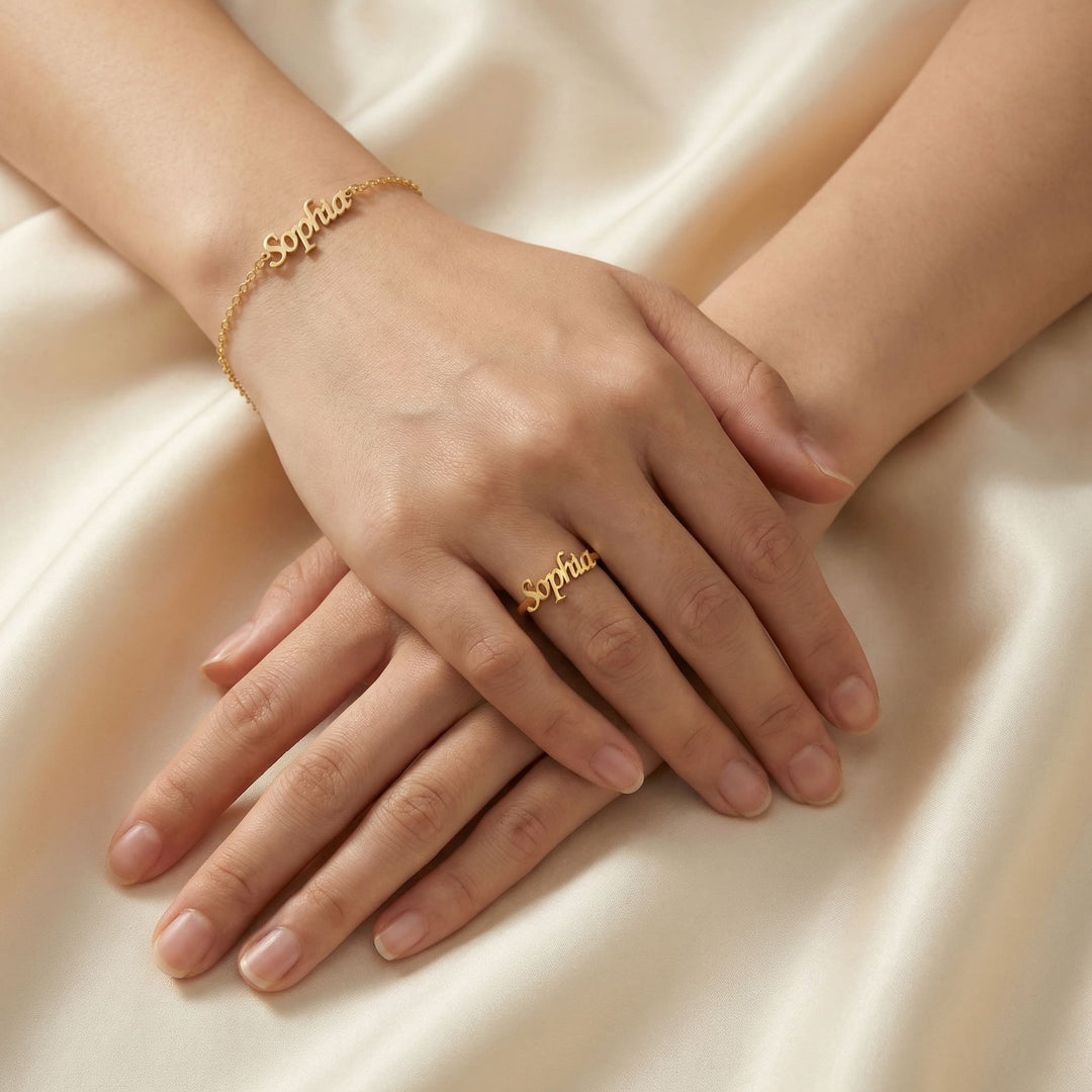 Woman's hands resting on cream satin fabric wearing matching gold custom name jewelry set featuring "Sophia" in cursive script, with a delicate name bracelet on the wrist and adjustable name ring on the finger
