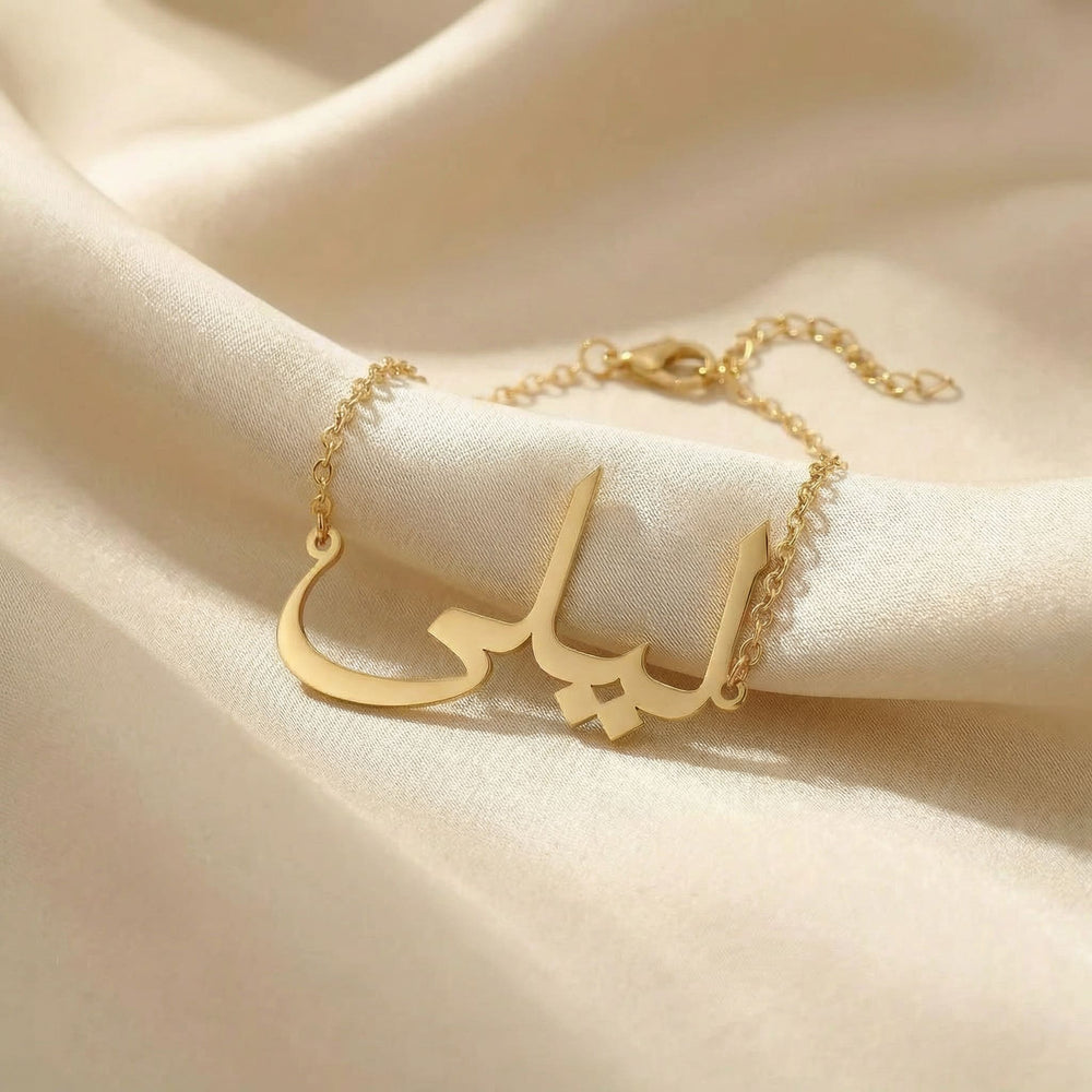 Custom Arabic name bracelet in gold featuring "ليلى" (Layla) in elegant Arabic calligraphy script with delicate chain and adjustable extender, displayed on cream satin fabric.