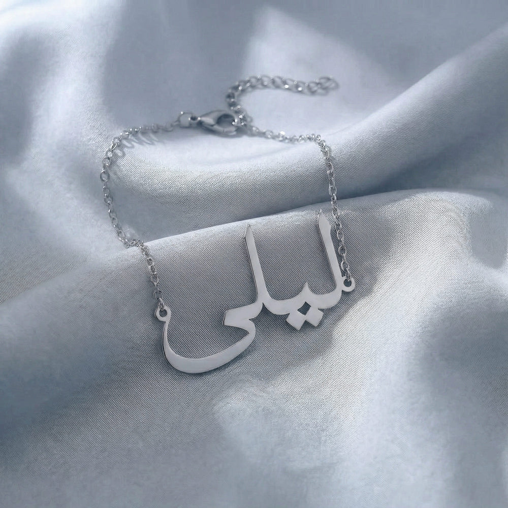 Custom Arabic name bracelet in silver featuring "ليلى" (Layla) in elegant Arabic calligraphy script with delicate chain and adjustable extender, displayed on light blue grey satin fabric.