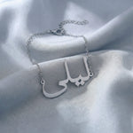 Custom Arabic name bracelet in silver featuring "ليلى" (Layla) in elegant Arabic calligraphy script with delicate chain and adjustable extender, displayed on light blue grey satin fabric.