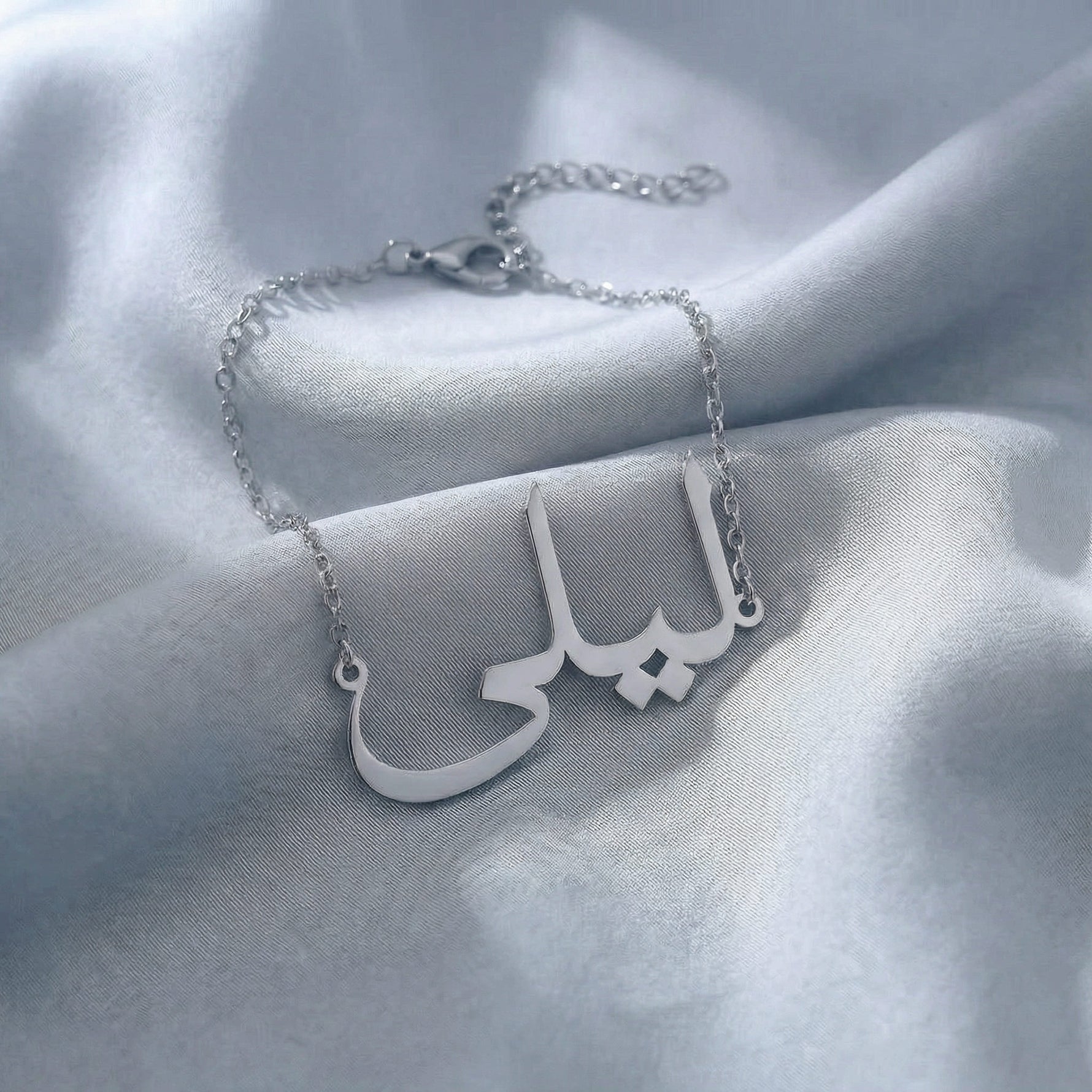 Custom Arabic name bracelet in silver featuring "ليلى" (Layla) in elegant Arabic calligraphy script with delicate chain and adjustable extender, displayed on light blue grey satin fabric.