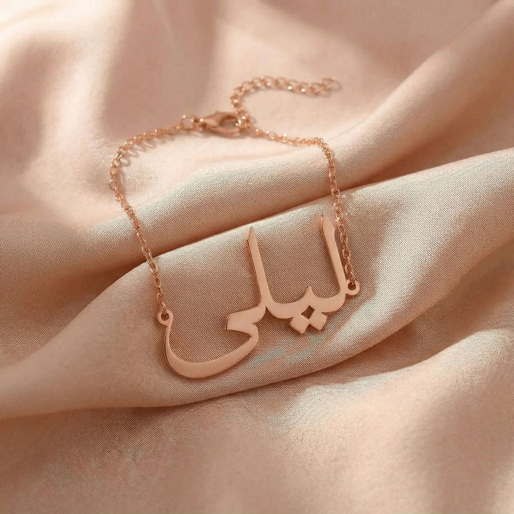 Custom Arabic name bracelet in rose gold featuring "ليلى" (Layla) in elegant Arabic calligraphy script with delicate chain and adjustable extender, displayed on blush pink satin fabric.