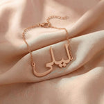 Custom Arabic name bracelet in rose gold featuring "ليلى" (Layla) in elegant Arabic calligraphy script with delicate chain and adjustable extender, displayed on blush pink satin fabric.