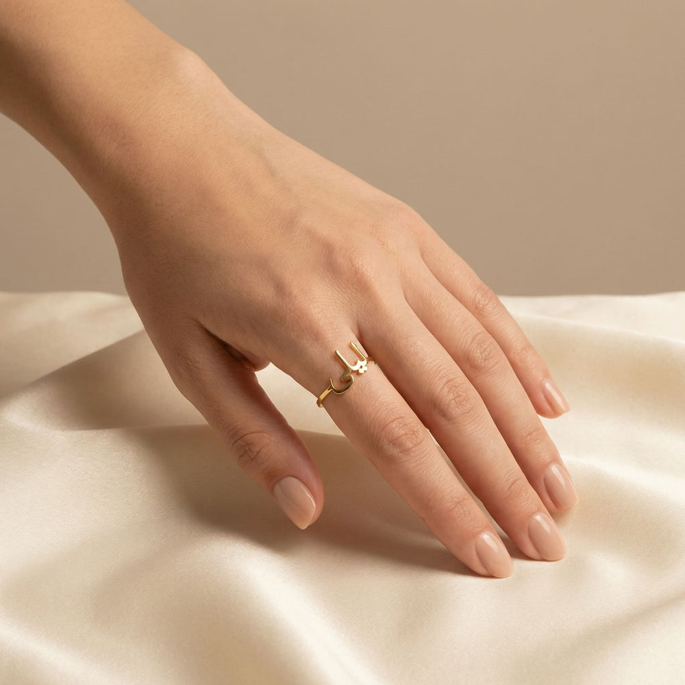 Woman's hand wearing a gold custom Arabic name ring on the middle finger, hand elegantly posed on cream satin fabric with nude nails.