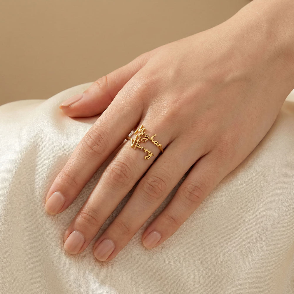Woman's hand wearing a gold custom couple name ring featuring "Meghan" and "Harry" in two rows of cursive script, hand resting softly on cream satin fabric.