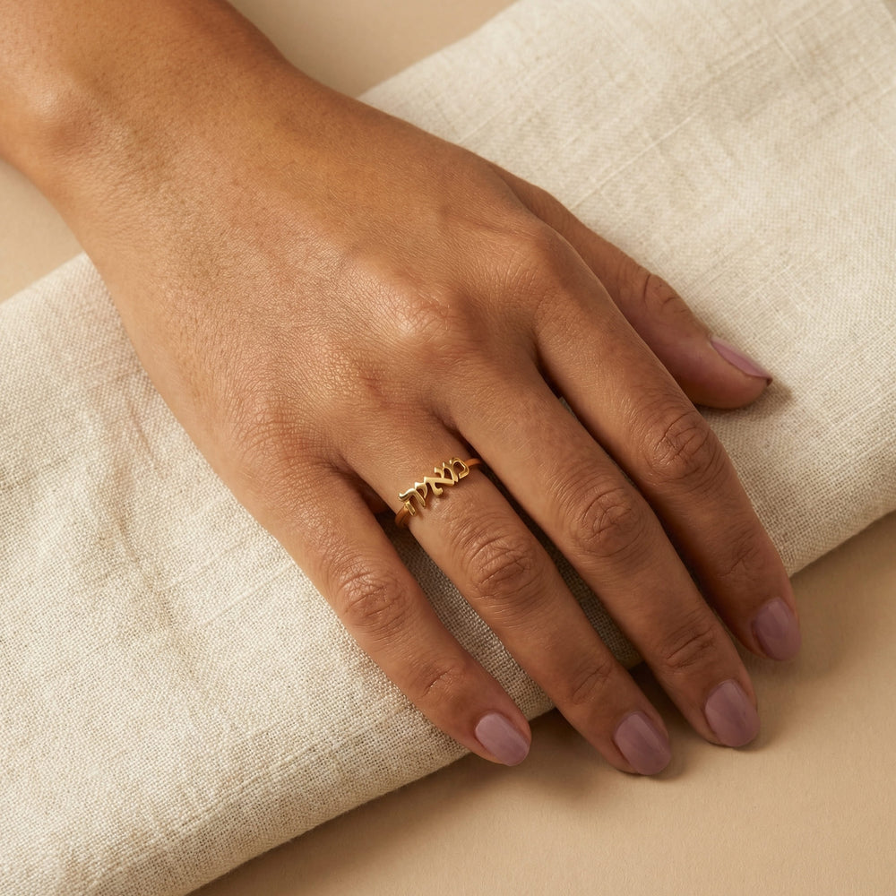 Woman's hand wearing a gold custom Hebrew name ring with "מאיה" (Maya) on the middle finger, hand resting on cream linen fabric with soft mauve nails.