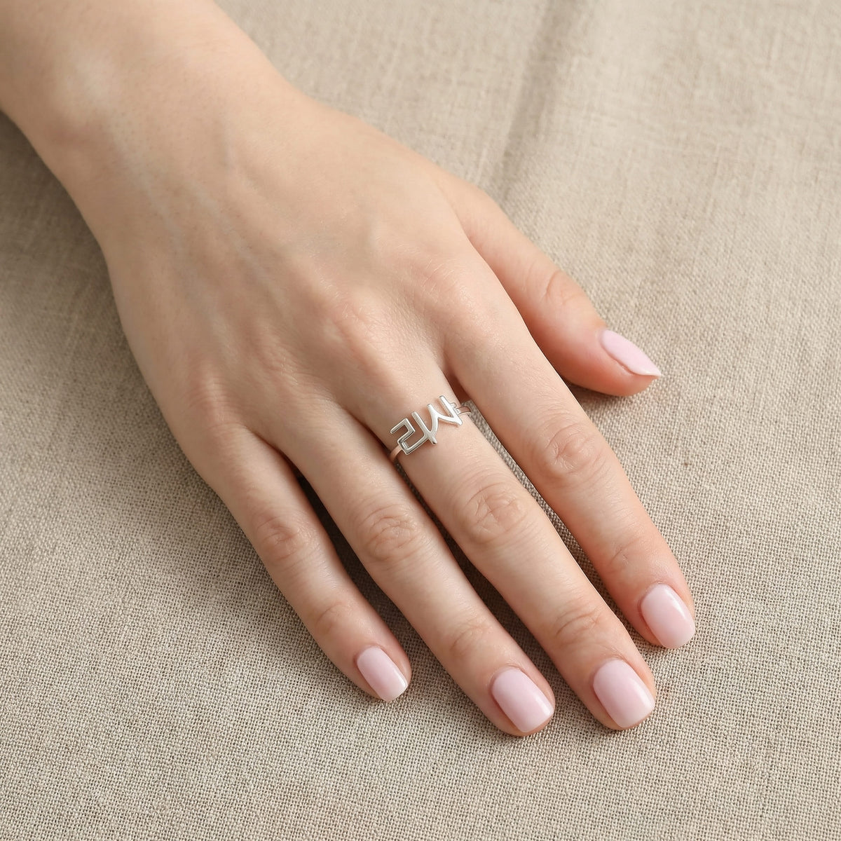 Woman's hand wearing a silver custom Korean Hangul name ring with "리사" (Lisa) on the middle finger, hand flat on beige linen fabric with soft pink nails.