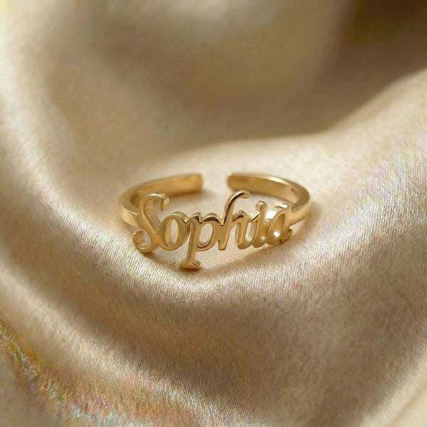 Custom name ring in gold with "Sophia" written in elegant cursive script, featuring an adjustable open band design, displayed on cream satin fabric.