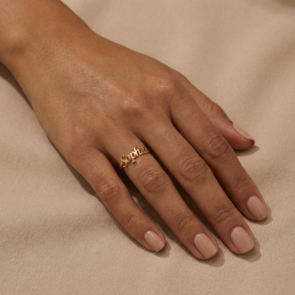 Woman's hand wearing a gold custom name ring with "Sophia" in cursive script on the middle finger, hand relaxed on beige satin fabric with nude nails.