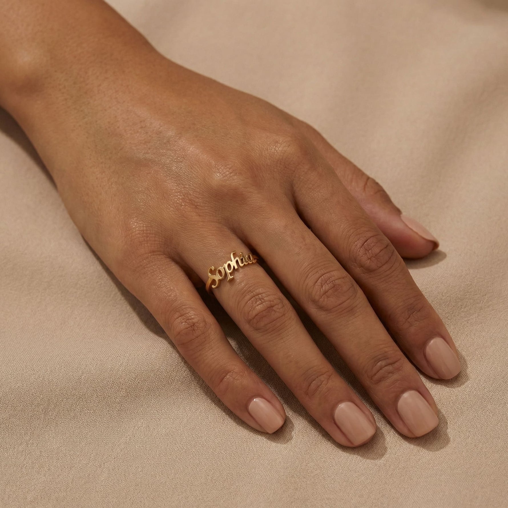 Woman's hand wearing a gold custom name ring with "Sophia" in cursive script on the middle finger, hand relaxed on beige satin fabric with nude nails.