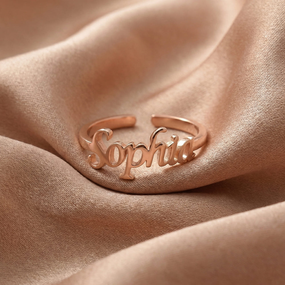 Custom name ring in rose gold with "Sophia" written in elegant cursive script, featuring an adjustable open band design, displayed on beige satin fabric.