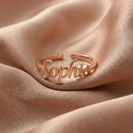Custom name ring in rose gold with "Sophia" written in elegant cursive script, featuring an adjustable open band design, displayed on beige satin fabric.