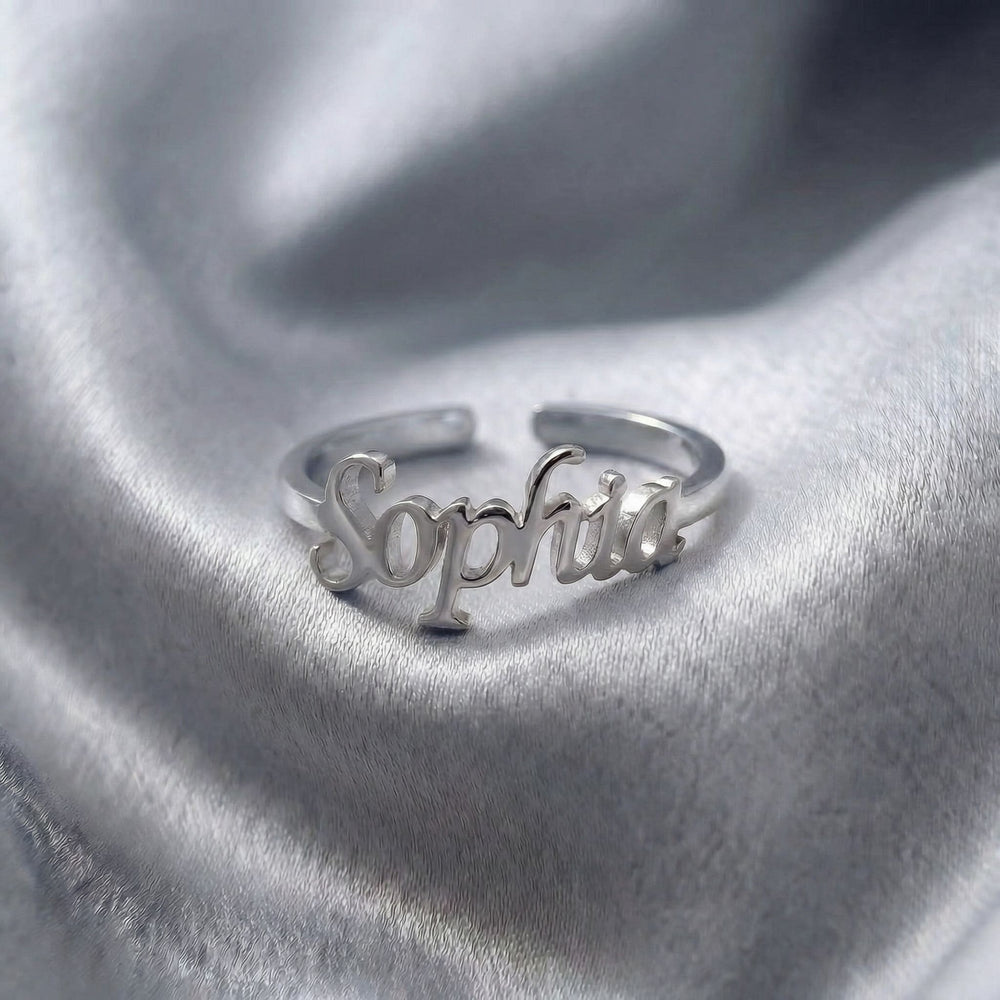 Custom name ring in silver with "Sophia" written in elegant cursive script, featuring an adjustable open band design, displayed on grey satin fabric.