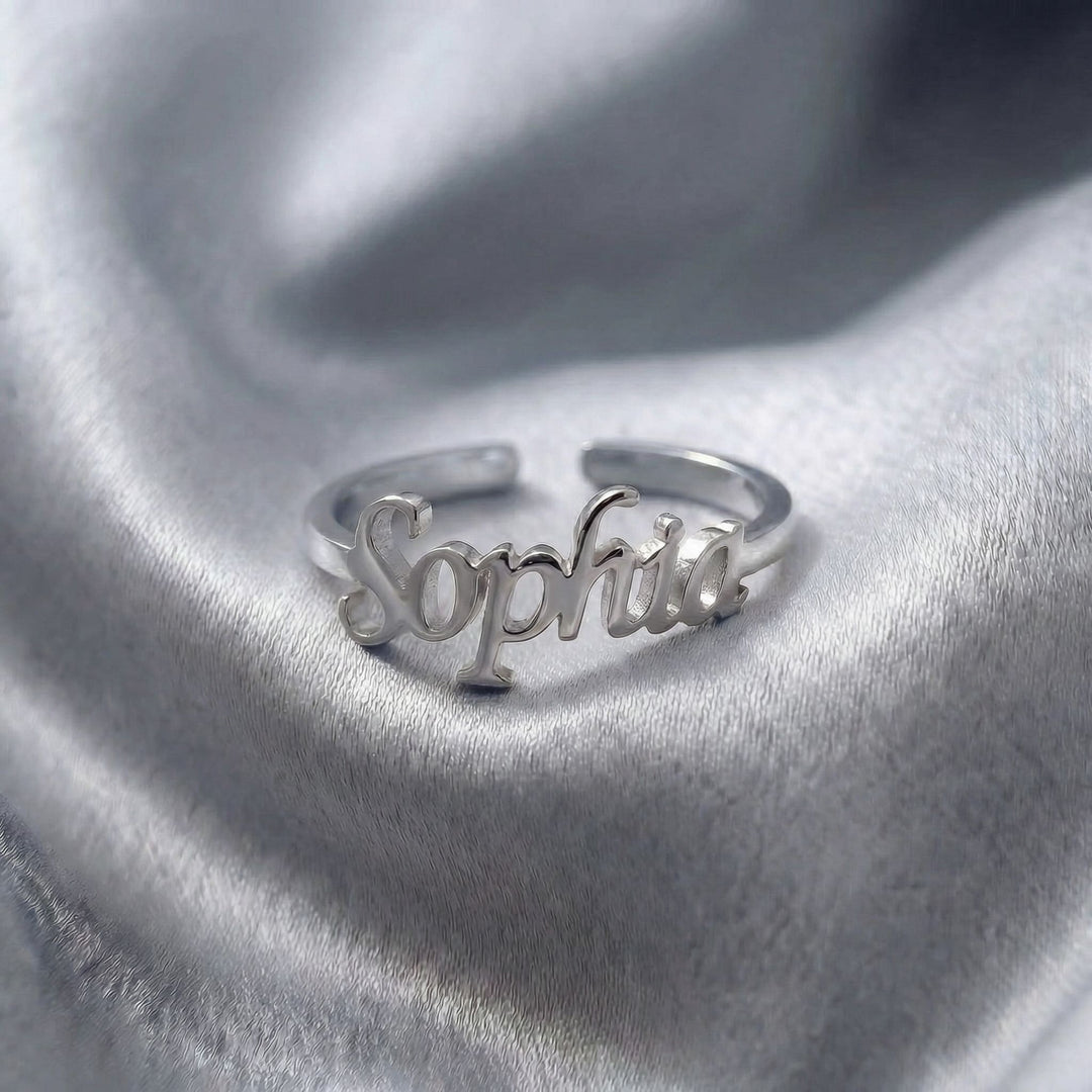 Custom name ring in silver with "Sophia" written in elegant cursive script, featuring an adjustable open band design, displayed on grey satin fabric.