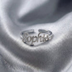 Custom name ring in silver with "Sophia" written in elegant cursive script, featuring an adjustable open band design, displayed on grey satin fabric.