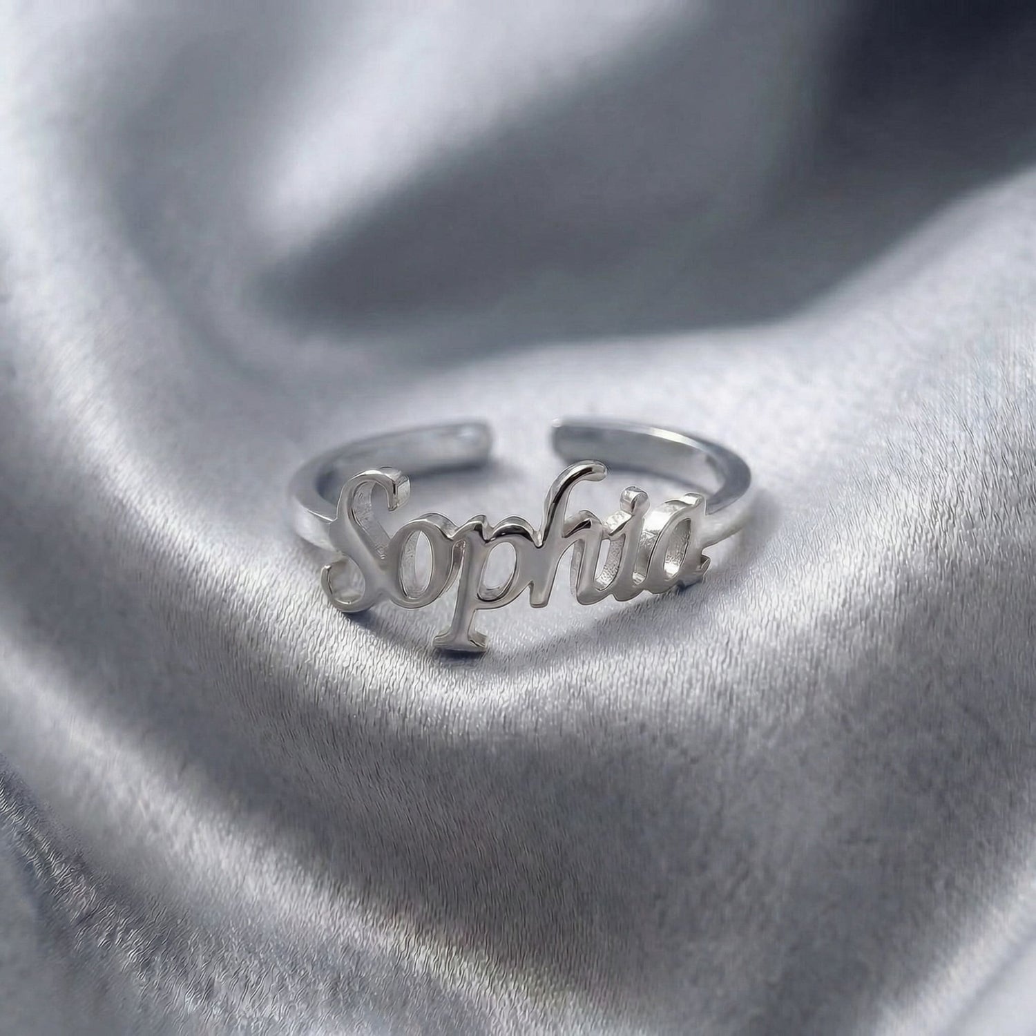 Custom name ring in silver with "Sophia" written in elegant cursive script, featuring an adjustable open band design, displayed on grey satin fabric.