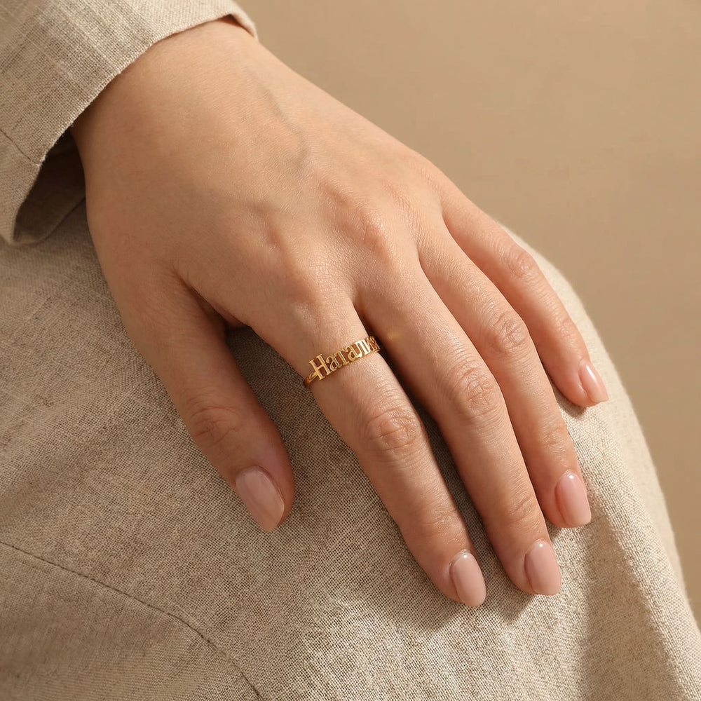 Woman's hand wearing a gold custom Cyrillic name ring with "Наталия" (Natalia) on the middle finger, hand resting on lap with beige linen clothing and nude pink nails.