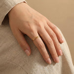 Woman's hand wearing a gold custom Cyrillic name ring with "Наталия" (Natalia) on the middle finger, hand resting on lap with beige linen clothing and nude pink nails.