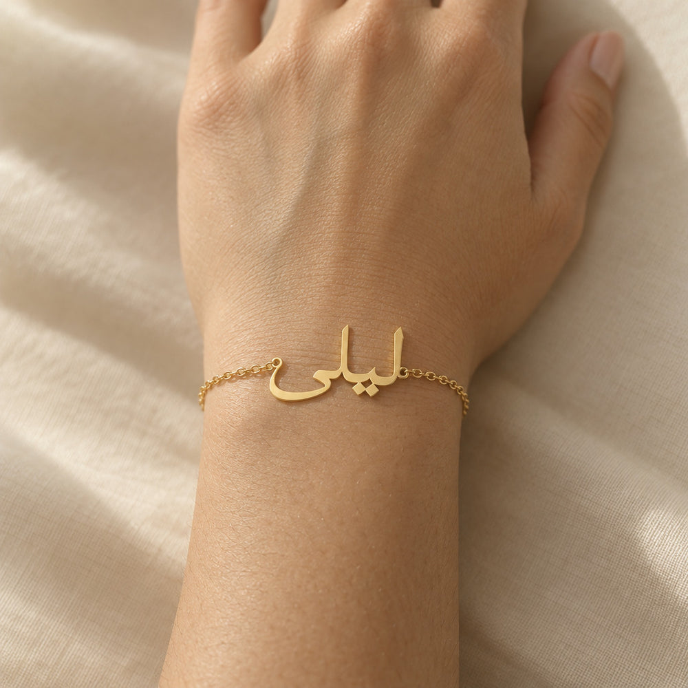 Close-up of a custom Arabic name bracelet in 18K gold plated finish worn on the wrist, the delicate nameplate reading "ليلى" (Layla) in cursive Arabic calligraphy against warm skin and cream fabric.