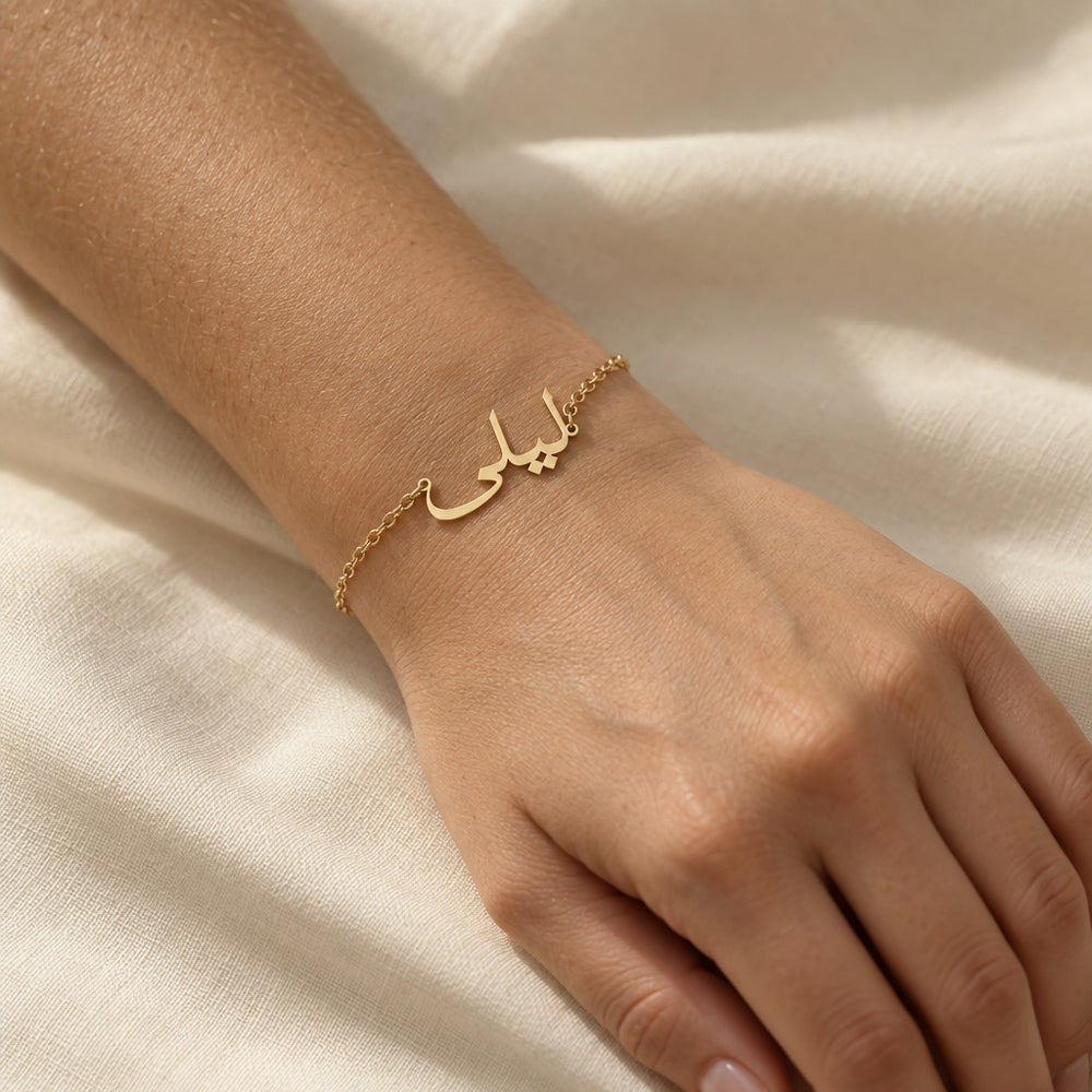 Woman's wrist wearing a custom Arabic name bracelet in 18K gold plated finish, the nameplate reading "ليلى" (Layla) in cursive Arabic script on a fine chain, hand resting on cream linen.