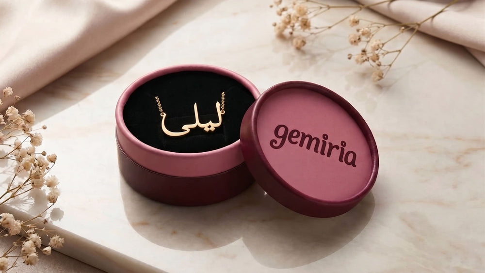 Burgundy-pink gift box with 'gemiria' branding with a Arabic name necklace inside on a marble surface with flowers.