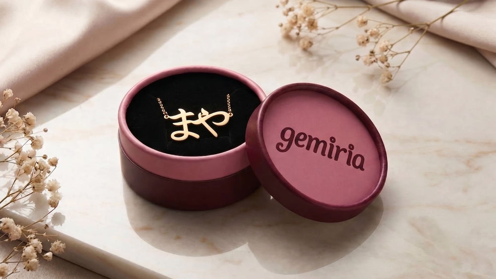 Burgundy-pink gift box with 'gemiria' branding with a Japanese name necklace inside on a marble surface with flowers.