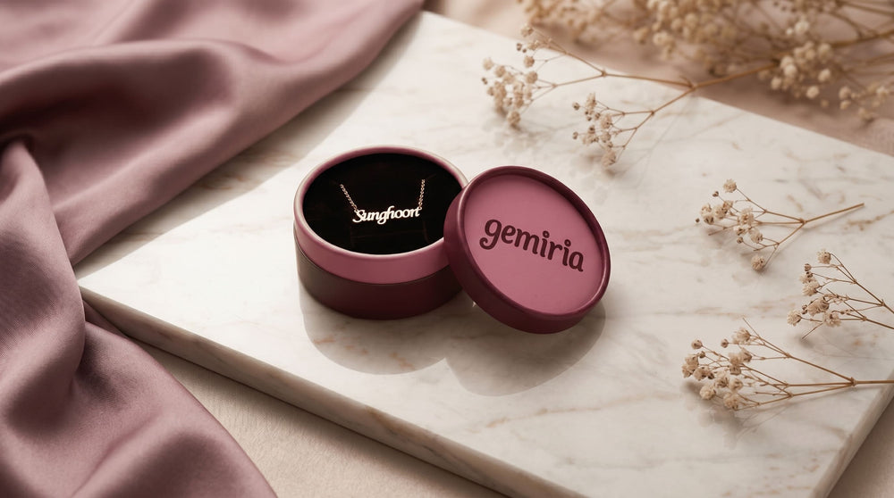 Burgundy-pink gift box with 'gemiria' branding with a 'Sunghoon' name necklace inside on a marble surface with flowers.