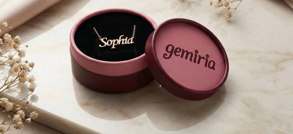 Open pink-burgundy Gemiria branded jewelry box with 'Sofia' name necklace inside, on a marble surface with flowers.