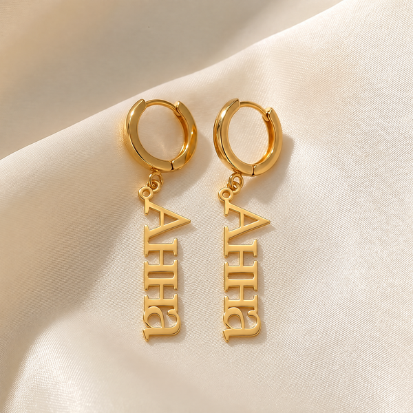Custom Cyrillic name drop earrings in 18K gold plating, a pair of huggie hoops with vertical name plates reading Анна, on ivory silk
