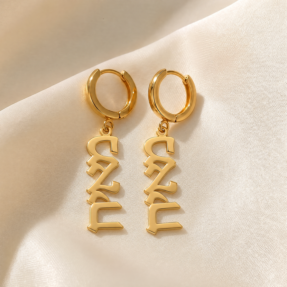 Custom Hebrew name drop earrings in 18K gold plating, a pair of huggie hoops with vertical name plates reading מאיה, on ivory silk